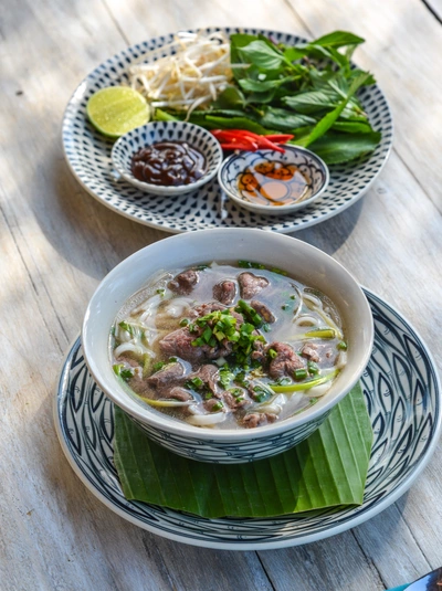 Ein Teller mit vietnamesischer Pho-Suppe, garniert mit frischem vietnamesischen Koriander (Coriandrum sativum) und anderen Kräutern, steht neben einer weiteren Platte mit Sprossen, Limetten und Chilis. Die Suppe enthält zarte Fleischscheiben und grüne Zwiebeln.