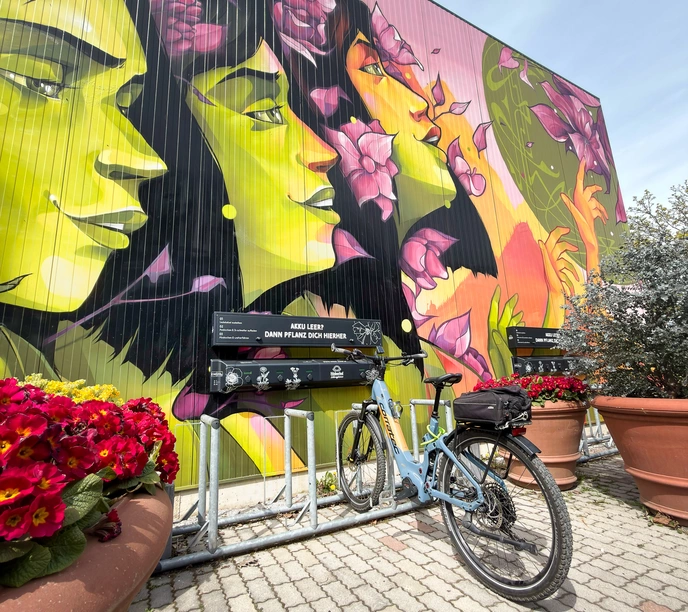 In diesem Bild kann man die kostenlos E-Bike Ladestation bei der Erlebnisgärtnerei Hödnerhof in Ebbs sehen. Es steht ein Rad beim laden dort und der Hintergrund ist bunt und mit Blumen versehen.
