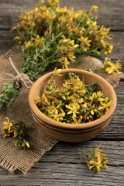 Johanniskraut (Hypericum perforatum) in getrockneter Form, mit gelben Blüten, die in einer Terrakottaschale liegen. Weitere getrocknete Zweige sind auf einem Jutestoff verteilt, was eine rustikale Atmosphäre erzeugt.