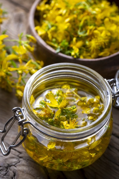 Johanniskraut (Hypericum perforatum) in einem Glasbehälter mit Öl, wobei die gelben Blüten im Öl schwimmen. Ein weiterer Holzschalenbehälter mit getrockneten Blüten ist ebenfalls im Bild sichtbar, was eine rustikale Stimmung erzeugt.