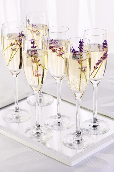 Sechs Sektgläser mit sprudelndem Getränk stehen auf einem weißen Tablett. Jedes Glas ist mit einem Zweig violetten Lavendels dekoriert. Die Szene ist hell beleuchtet, mit einem weißen Hintergrund, der die elegante Präsentation betont.
