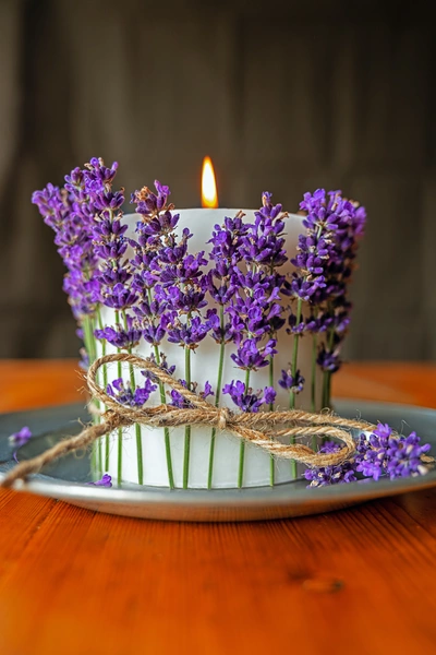 Eine brennende weiße Kerze, umwickelt mit Lavendelzweigen und einer rustikalen Juteschnur, steht auf einem grauen Teller auf einem Holztisch. Der Hintergrund ist dunkel und unscharf, wodurch die violetten Lavendelblüten und das warme Kerzenlicht hervorgehoben werden.