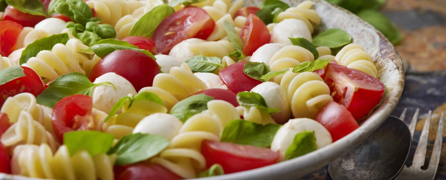 Eine Schüssel mit Nudelsalat, der mit Mini-Basilikum (Ocimum basilicum) dekoriert ist. Zwischen den Nudeln sind Kirschtomaten und kleine Mozzarellakugeln verteilt, die für ein frisches, mediterranes Aussehen sorgen. Im Hintergrund sind eine Gabel und ein Löffel zu sehen.