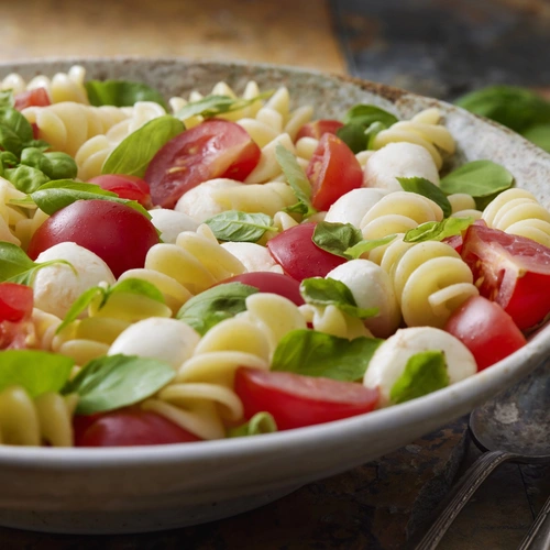 Eine Schüssel mit Nudelsalat, der mit Mini-Basilikum (Ocimum basilicum) dekoriert ist. Zwischen den Nudeln sind Kirschtomaten und kleine Mozzarellakugeln verteilt, die für ein frisches, mediterranes Aussehen sorgen. Im Hintergrund sind eine Gabel und ein Löffel zu sehen.