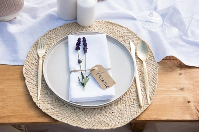 Ein gedeckter Holztisch mit einem runden Teller auf einem geflochtenen Platzset. Auf der weißen Serviette liegt ein Lavendelzweig, gebunden mit einer Schnur und einem Namensschild. Goldfarbenes Besteck, weiße Kerzen und Gläser vervollständigen das elegante Setting.