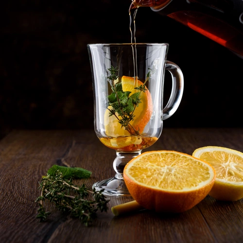 Heißer Tee wird in ein Glas mit Orangenscheiben und frischem Orangen-Thymian gegossen. Daneben liegen auf einer dunklen Holzoberfläche frische Orangenhälften und Thymianzweige.