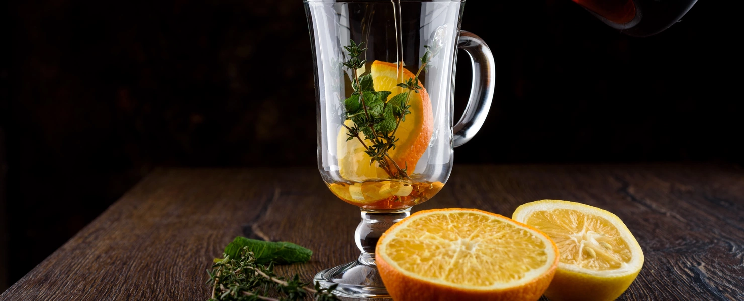 Heißer Tee wird in ein Glas mit Orangenscheiben und frischem Orangen-Thymian gegossen. Daneben liegen auf einer dunklen Holzoberfläche frische Orangenhälften und Thymianzweige.