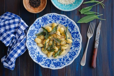 Ein Teller mit Gnocchi und knusprig gebratenem Salbei auf einem blauen Blumenmuster-Teller. Daneben liegen Besteck, frische Salbeiblätter, Pfefferkörner und eine Schüssel mit geriebenem Käse auf einem dunklen Holztisch. Eine karierte Serviette ist gefaltet daneben.