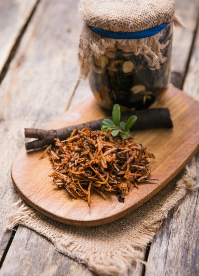 Getrocknete Wurzelstücke von Echtem Beinwell auf einem Holzbrett, daneben frische Wurzelstücke und ein kleines grünes Blatt. Im Hintergrund ein Glas mit dunkler Flüssigkeit, mit Stoff und Kordel abgedeckt, auf einer rustikalen Holzoberfläche.