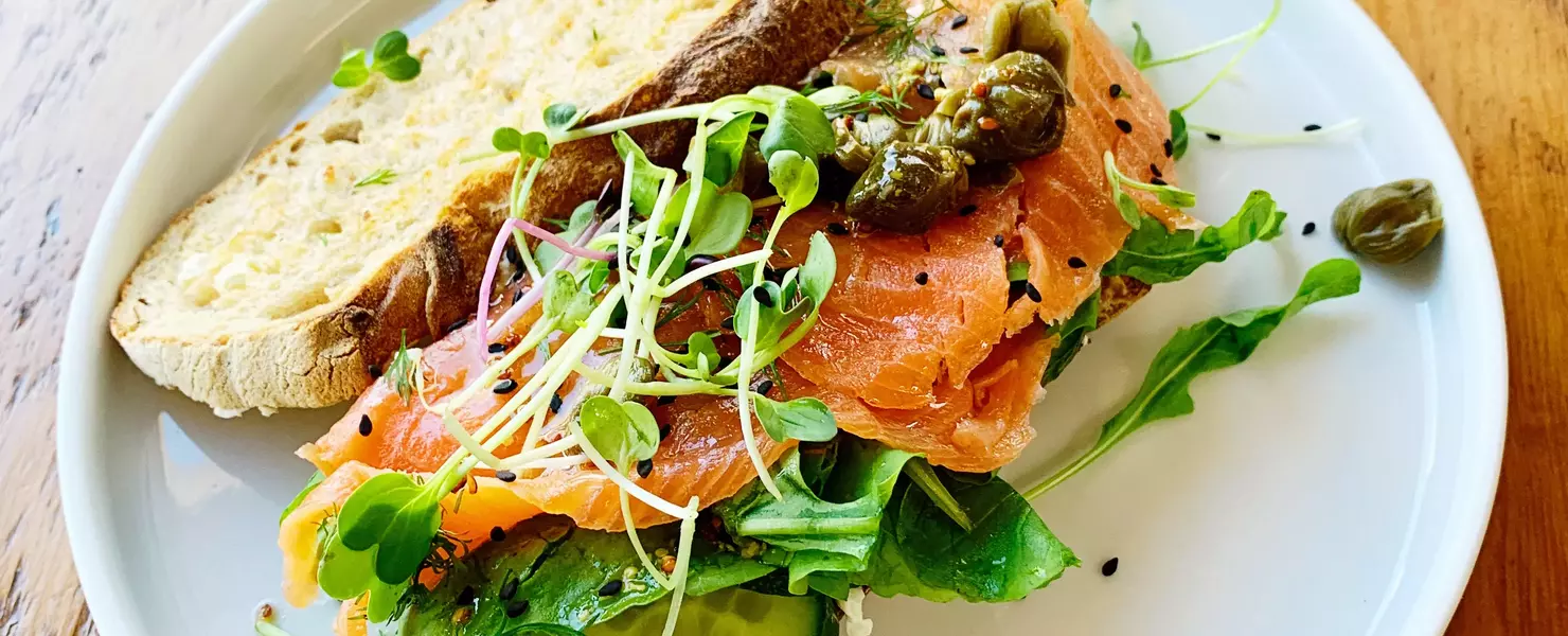 Ein Sandwich mit geräuchertem Lachs, frischer Brunnenkresse (Nasturtium officinale), Gurkenscheiben und Kapern. Das Sandwich ist auf einem weißen Teller angerichtet und wirkt frisch und einladend.