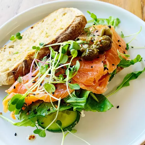 Ein Sandwich mit geräuchertem Lachs, frischer Brunnenkresse (Nasturtium officinale), Gurkenscheiben und Kapern. Das Sandwich ist auf einem weißen Teller angerichtet und wirkt frisch und einladend.