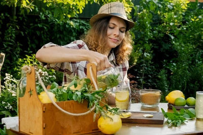Eine Frau mit lockigem Haar und Strohhut sitzt an einem Tisch im Garten, bereitet ein Getränk mit frischer Hugo-Minze, Zitrusfrüchten und einer Karaffe zu, umgeben von natürlicher Vegetation.
