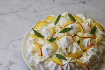 Eine Pavlova-Torte mit Baiser, Schlagsahne, frischen Pfirsichscheiben und Zitronenverbene-Blättern auf einem weißen Teller. Der Hintergrund ist eine Marmoroberfläche.