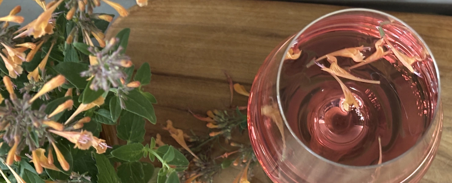 Eine Limonadenpflanze (Aloysia citrodora) mit orangefarbenen Blüten steht neben einem Glas mit pinkfarbener Flüssigkeit, in dem Blütenblätter schwimmen. Das Bild zeigt die Pflanze in der Nähe des Glases auf einem Holztisch.