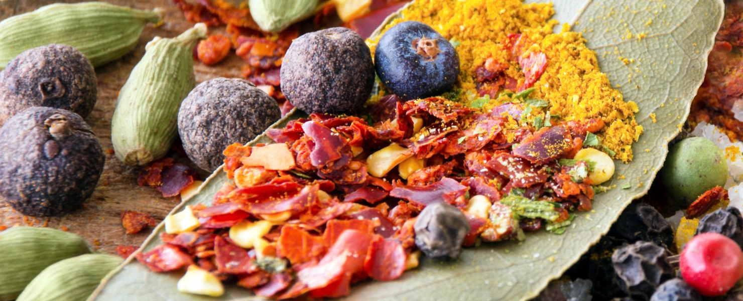 Verschiedene Gewürze, darunter Lorbeerblätter, Gewürz-Thymian, Currypulver, Wacholderbeeren und bunte Pfefferkörner, liegen dekorativ arrangiert auf einer Oberfläche. Die leuchtenden Farben der Gewürze betonen ihre Vielfalt und Frische.