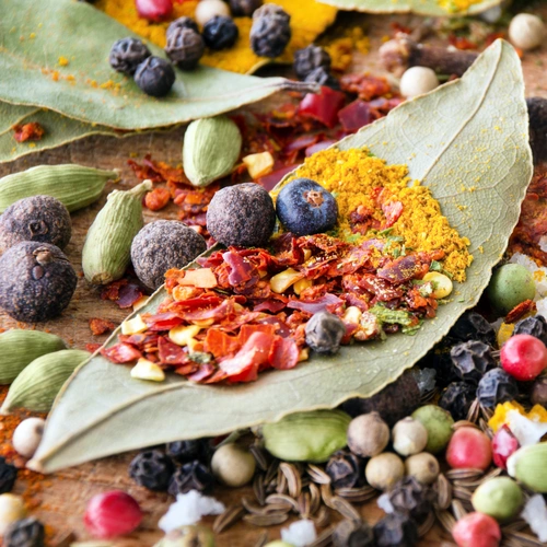 Verschiedene Gewürze, darunter Lorbeerblätter, Gewürz-Thymian, Currypulver, Wacholderbeeren und bunte Pfefferkörner, liegen dekorativ arrangiert auf einer Oberfläche. Die leuchtenden Farben der Gewürze betonen ihre Vielfalt und Frische.