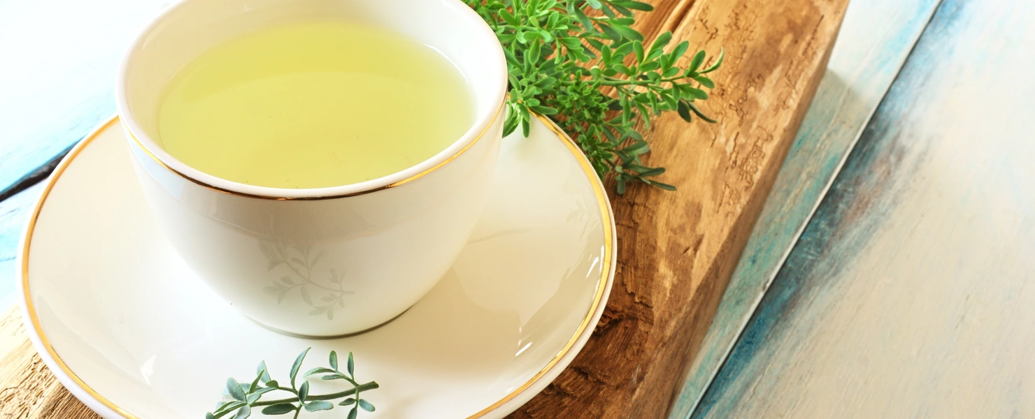 Eine weiße Teetasse mit goldener Verzierung, gefüllt mit gelblichem Weinrautentee, steht auf einer Untertasse auf einem rustikalen Holzbrett. Daneben liegen frische Zweige der Weinraute.