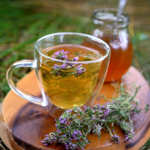 Ein Glas mit Kräutertee, in dem Quendel-Arzneithymian schwimmt, steht auf einem Holzbrett im Freien. Daneben liegen frische Thymianzweige und ein Glas Honig mit Löffel. Der Hintergrund ist unscharf und zeigt grünes Gras.