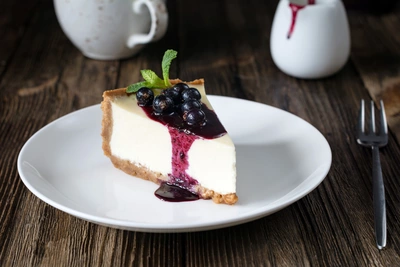 Ein Stück Käsekuchen mit Johannisbeer-Sauce, dekoriert mit frischen Johannisbeeren und Minzblättern auf einem weißen Teller, daneben eine Gabel. Im Hintergrund ein Krug und eine Tasse auf dunklem Holztisch.