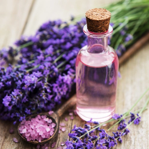 Ein kleines Glasfläschchen mit rosafarbenem Lavendelöl steht auf einer rustikalen Holzoberfläche. Daneben liegen frische Lavendelblüten, eine Holzlöffel mit rosa Badesalz und verstreute Salzkristalle. Die Szene wirkt natürlich und entspannend.