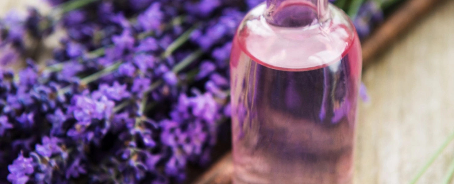 Ein kleines Glasfläschchen mit rosafarbenem Lavendelöl steht auf einer rustikalen Holzoberfläche. Daneben liegen frische Lavendelblüten, eine Holzlöffel mit rosa Badesalz und verstreute Salzkristalle. Die Szene wirkt natürlich und entspannend.