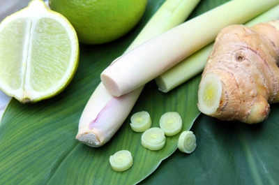Frische Zutaten auf einem grünen Bananenblatt: Ganze und geschnittene Zitronengrasstängel mit blassgrüner Farbe, daneben eine halbe und eine ganze Limette sowie ein Stück Ingwer mit heller, leicht strukturierter Schale.