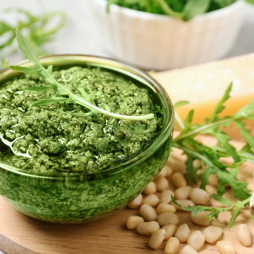 Grünes Rucola-Pesto in einer Glasschüssel auf einem Holzbrett, dekoriert mit frischem Rucola. Daneben liegen Pinienkerne, eine Knoblauchzehe und ein Stück Parmesan. Im Hintergrund eine Schüssel mit Rucola. Heller Untergrund, leicht unscharfer Hintergrund.