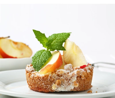 Ein Dessert aus Streuselkuchen mit Apfelscheiben und einem frischen Apfelminzblatt als Garnitur, serviert auf einem weißen Teller vor einem hellen Hintergrund.