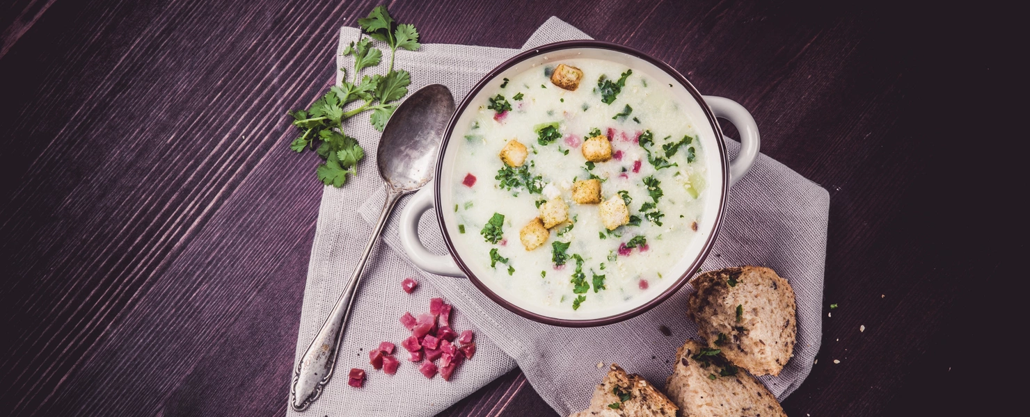 Eine Schale cremige Suppe, garniert mit Koriander, Croutons und Granatapfelkernen, auf einem dunklen Holztisch. Daneben ein Löffel, frische Korianderzweige und aufgeschnittenes Brot auf einem Leinentuch.