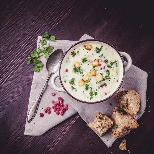 Eine Schale cremige Suppe, garniert mit Koriander, Croutons und Granatapfelkernen, auf einem dunklen Holztisch. Daneben ein Löffel, frische Korianderzweige und aufgeschnittenes Brot auf einem Leinentuch.