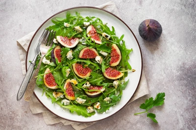 Frischer Rucola-Salat mit Feigenscheiben, Ziegenkäse und Walnüssen auf einem weißen Teller mit braunem Rand. Daneben eine Gabel, eine Serviette und eine ganze Feige auf hellem Steinuntergrund.