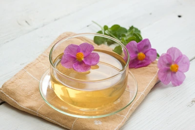 Eine Tasse goldener Zistrosentee mit einer pinken Zistrosenblüte darauf, stehend auf einer beigen Serviette. Daneben liegen weitere frische Zistrosenblüten und grüne Blätter auf einem weißen Holztisch.