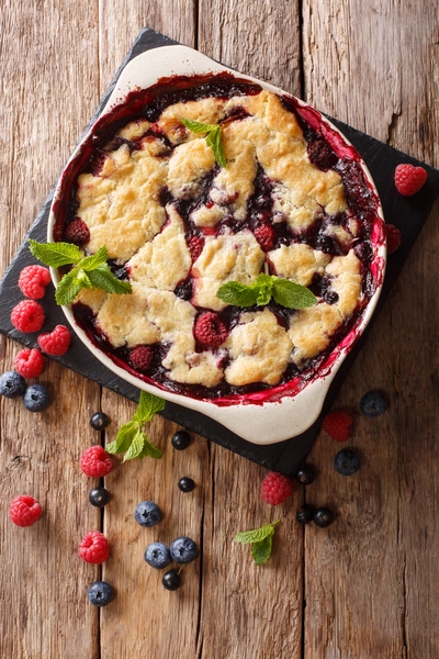 Ein frischer Beeren-Crumble in einer quadratischen Backform auf dunklem Holzuntergrund, dekoriert mit Johannisbeeren, Blaubeeren, Himbeeren und Minzblättern. Einige Beeren liegen verstreut um die Form.