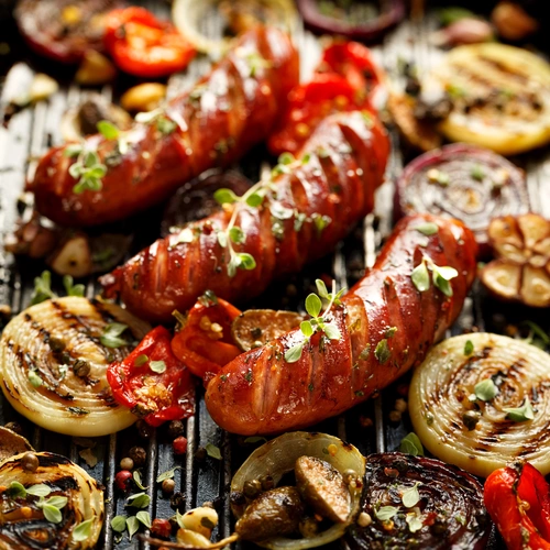 Gegrillte Würstchen mit Gemüse wie Zwiebeln, Tomaten und Knoblauch, garniert mit frischem einjährigem Majoran (Origanum marjorana). Das Bild zeigt eine appetitliche, rustikale Mahlzeit mit einer Vielzahl von Aromen.