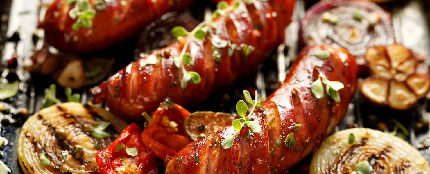 Gegrillte Würstchen mit Gemüse wie Zwiebeln, Tomaten und Knoblauch, garniert mit frischem einjährigem Majoran (Origanum marjorana). Das Bild zeigt eine appetitliche, rustikale Mahlzeit mit einer Vielzahl von Aromen.