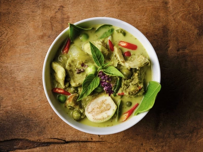 Eine Schüssel mit thailändischem Curry, garniert mit frischen Blättern von Thai-Basilikum (Ocimum basilicum var. thyrsiflora), roten Chilischoten und grünen Erbsen. Das Curry hat eine grüne Farbe und enthält zarte Fleischstücke.