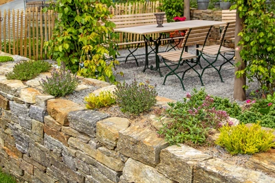 Ein Steingarten mit echtem Thymian und anderen Kräutern wächst auf einer Trockenmauer, im Hintergrund eine gemütliche Sitzgruppe mit Holztisch und Bänken unter einer begrünten Pergola.