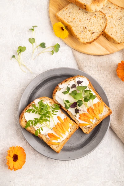 Zwei Scheiben Brot mit Frischkäse, verziert mit Ringelblumenblättern und frischen Kräutern, auf einem grauen Teller. Um das Essen sind orangefarbene Ringelblumen und Sprossen verstreut.
