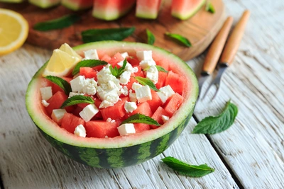 Eine halbe Wassermelone, gefüllt mit würfelförmigen Melonenstücken, garniert mit frischen Minzblättern und Feta-Käse. Im Hintergrund sind Zitronenscheiben, Minzblätter und ein Holzbrett mit Messern auf einer hellen Holzoberfläche zu sehen.