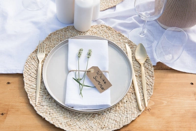 Ein gedeckter Holztisch mit einem runden, geflochtenen Platzset. Darauf ein grauer Teller mit weißer Serviette, verziert mit zwei weißen Lavendelzweigen und einem Anhänger mit der Aufschrift „White Summer“. Daneben goldfarbenes Besteck, Kerzen und Gläser.