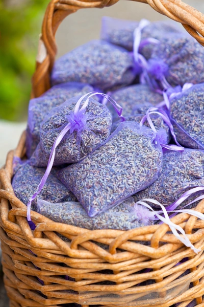 Ein geflochtener Korb, gefüllt mit kleinen Lavendelsäckchen aus transparentem Stoff. Jedes Säckchen ist mit getrocknetem Lavendel gefüllt und mit einem lila Satinband verschlossen. Der Hintergrund ist leicht unscharf und zeigt grünes Laub.