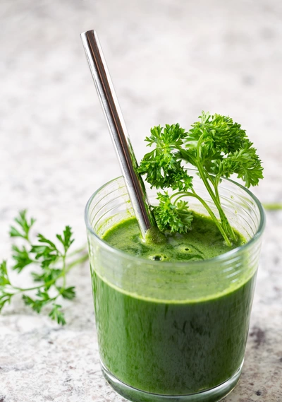 Ein Glas grüner Smoothie, garniert mit frischer Krauser Petersilie (Petroselinum crispum). Der Smoothie ist leuchtend grün und das Glas enthält einen Metallstrohhalm, was dem Bild eine gesunde, moderne Ausstrahlung verleiht.