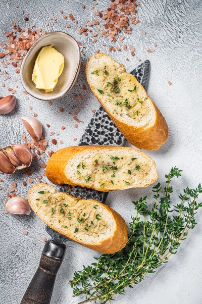 Ein knuspriges Brot mit einer Kräuterbutter, die mit frischem, grünen Thymian und Knoblauch belegt ist, liegt auf einer grauen Oberfläche, umgeben von Salzflocken, Knoblauchzehen und einem Messer.