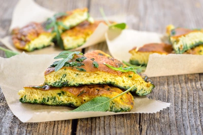 Stücke von goldbrauner Frittata mit Salbeiblättern liegen auf Backpapier auf einem rustikalen Holztisch. Im Hintergrund sind weitere Stücke der Frittata zu sehen.