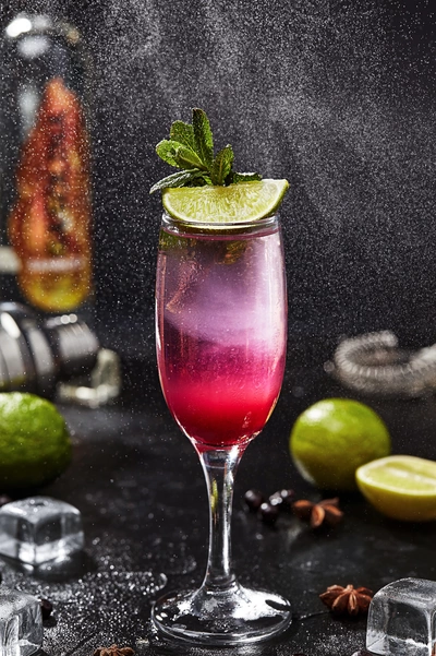 Ein elegantes Glas mit einem roten Johannisbeer-Minze-Drink, garniert mit Minzblättern und einer Limettenscheibe, auf einem dunklen Hintergrund mit dekorativen Limetten und Eiswürfeln.