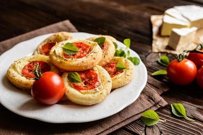 Runde Blätterteiggebäcke auf einem weißen Teller, belegt mit Tomatenscheiben, Käse und frischen Oregano-Blättern, dekoriert mit ganzen Tomaten und Käse im Hintergrund auf einem dunklen Holztisch.