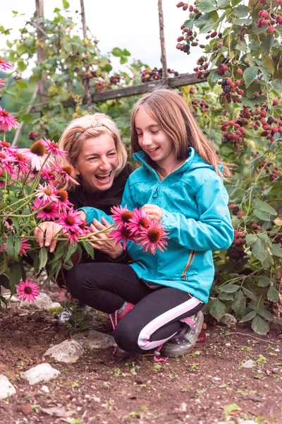 Das Bild zeigt zwei Personen, eine Erwachsene und ein Kind, die zusammen Arznei-Sonnenhut (Echinacea)-Blüten pflücken. Sie stehen in einem Garten, umgeben von üppigem Grün und blühenden Pflanzen, und lächeln während sie mit den Blumen interagieren.