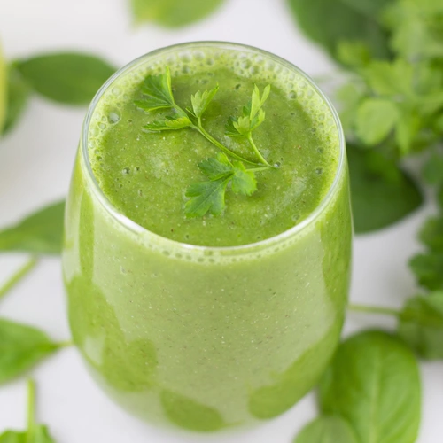 Ein Glas grüner Smoothie, garniert mit frischen Blättern der Glatten Petersilie (Petroselinum crispum). Der Smoothie enthält vermutlich eine Mischung aus grünen Zutaten wie Obst und Gemüse, und das Bild zeigt eine erfrischende, gesunde Getränkezubereitung.