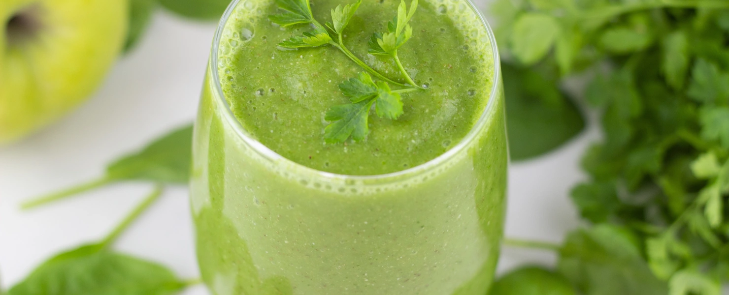 Ein Glas grüner Smoothie, garniert mit frischen Blättern der Glatten Petersilie (Petroselinum crispum). Der Smoothie enthält vermutlich eine Mischung aus grünen Zutaten wie Obst und Gemüse, und das Bild zeigt eine erfrischende, gesunde Getränkezubereitung.