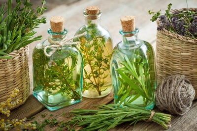 Drei Flaschen mit Kräuterölen, die mit verschiedenen Kräutern wie Bergbohnenkraut (Satureja montana), Rosmarin und Thymian gefüllt sind, stehen auf einem rustikalen Holztisch. Die Flaschen sind mit Korken verschlossen und umgeben von Kräuterzweigen und einer Rolle Jutegarn.